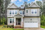 A two-story modern suburban house at 25 Cackling Way, Zebulon, with gray siding, white trim, and a brick base. It features a covered front porch, double garage, and is surrounded by tall pine trees and a green lawn.