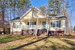Charming single-story beige house at 103 Pueblo Drive, Louisburg, featuring a green door, white porch railings, front steps, and a sloped roof, nestled among trees and a lush lawn on a sunny day.