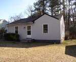 A small, single-story house at 105 S Elizabeth Street in Henderson features white siding, a dark shingle roof, and a brick chimney; two front windows frame the central door with small steps, set amid a grassy yard with trees and shrubs.