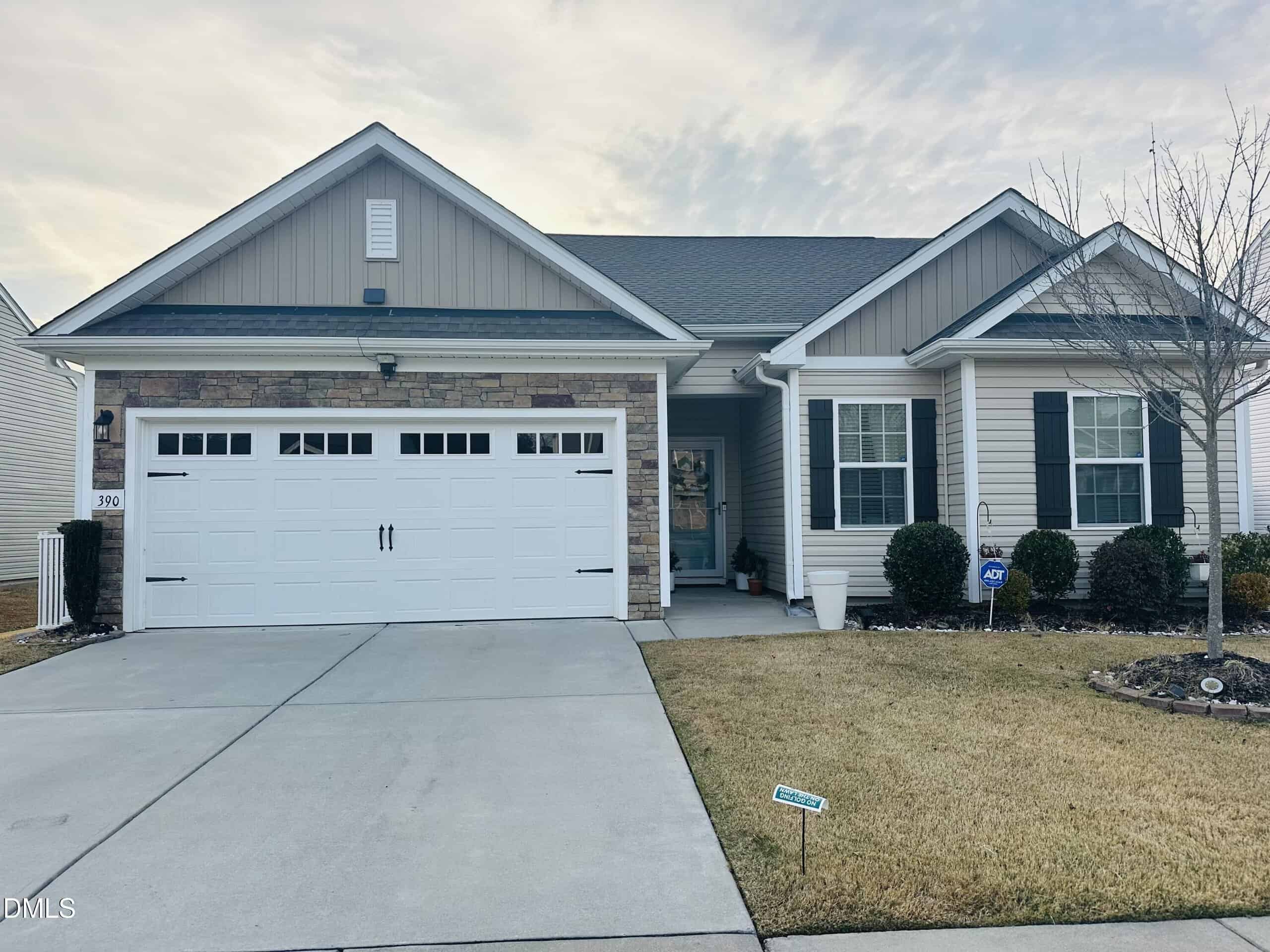 Charming single-story suburban house in Youngsville at 390 Legacy Drive, featuring a double garage, beige siding, stone accents, and dark shutters. The front yard boasts trimmed bushes, a small tree, and a well-maintained lawn with a curved driveway.