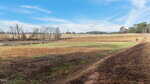A wide, open field with patches of green and brown grass lies off 0 Bennett Perry Road, Louisburg. Scattered trees dot the land, with a small pond beneath a bright blue sky. A rural road curves along the right side of the image.