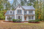 A two-story white house with gray shutters and roof at 3333 Keighley Forest Drive, Wake Forest, is surrounded by trees. It features a bay window, a welcoming front porch, wooden door, and a curved path bordered by shrubs and fresh mulch.