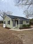 A small, single-story green house with black shutters and a black roof sits on a tidy lot at 317 Brewer Road in Louisburg, featuring a covered front porch, shrubs, propane tank, bare trees, and brown grass.