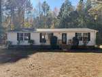 A single-story white house with black shutters and a small front porch sits at 192 Mum Lane in Henderson, set before tall pine trees and surrounded by a yard covered in pine needles.