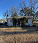 A small, single-story house at 501 Sunset Avenue, Oxford, with light-colored siding, red shutters, and a red front door, is surrounded by neatly trimmed bushes and trees on a grassy lawn under a clear blue sky.