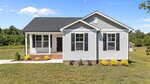 Charming single-story white house at 140 Brookhaven Drive, Spring Hope, with black shutters, a covered front porch, and a well-kept lawn. Small shrubs and yellow plants line the front, framed by trees under a clear blue sky.