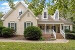 A beige two-story house with green shutters sits at 65 Coral Ridge Circle in Franklinton, featuring a covered front porch, white railings, brick steps, and a walkway surrounded by lush bushes and trees under a partly cloudy sky.
