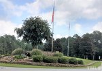 A landscaped stone entrance at 131 Mohawk Drive in Louisburg features a "Lake Royale" sign, vibrant flower beds, a large tree, and two flagpoles with American and North Carolina flags, all set among lush greenery under a partly cloudy sky.