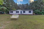 A single-story white house with dark shutters sits on a large grassy lawn at 110 Castlebury Lane, Franklinton, featuring a white decorative bench in front and surrounded by tall trees with dense green foliage under a partly cloudy sky.