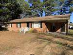 A charming one-story brick house at 41 Tant Road, Zebulon, features a gabled roof, white trim, and a small front porch with railings. The yard is blanketed in pine needles and surrounded by tall trees, with an attached carport on the right.