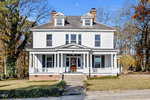 A large, two-story white house at 105 S Elm Street, Louisburg, features a wide front porch with columns, brick foundation, and two chimneys. The home sits on a grassy lot with trees and a concrete sidewalk in front.