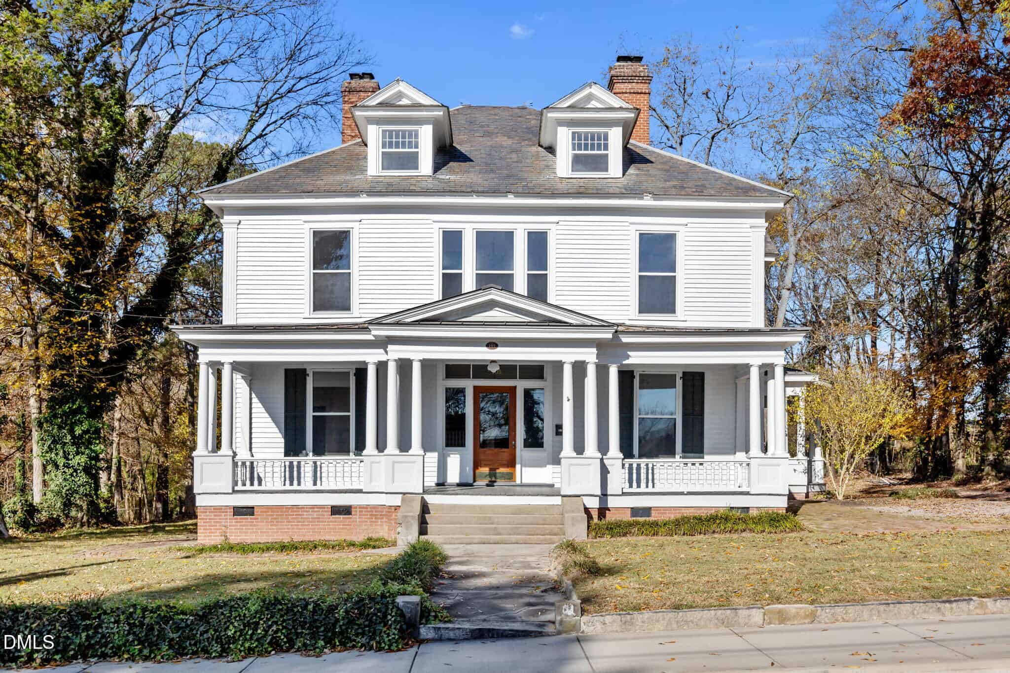A large, two-story white house at 105 S Elm Street, Louisburg, features a wide front porch with columns, brick foundation, and two chimneys. The home sits on a grassy lot with trees and a concrete sidewalk in front.