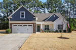 Charming single-story suburban house at 200 White Ash Lane, Youngsville, with gray siding and stone accents, a two-car garage, small front porch, manicured lawn, American flag on the right, and tall pine trees under a clear sky.