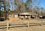 A one-story brick house with a front porch, brown roof, and black shutters sits behind a wooden fence at 121 Cokesbury Court. Leafless trees and a grassy yard surround this Henderson home under a clear blue sky.