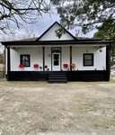 A white house with black trim at 332 Chavasse Avenue in Henderson features a covered front porch with two red chairs, potted plants, and three steps leading up from a wide concrete driveway. Trees border the yard under a cloudy sky.