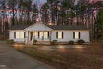 A white single-story house at 1739 Cobblestone Drive in Creedmoor features a front porch, black shutters, and a driveway, nestled among tall trees and beautifully illuminated by exterior lights at dusk.