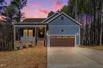 A blue single-story house at 128 Wichita Way in Louisburg features a front porch, brown double garage doors, and a concrete driveway, set against tall trees and a purple-pink sunset sky.