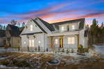 A modern white house with a dark roof and front porch, illuminated warmly at dusk at 115 Cherry Bark Drive, Youngsville. Snow blankets the ground and roof beneath a colorful sunset sky, framed by trees in the background.