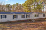A long, single-story manufactured home with white siding, dark blue shutters, and a brick foundation stands in front of a wooded area with tall trees and a clear sky. Steps lead to the front door.