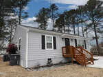 A small, single-story gray house with black shutters and a wooden front porch sits on a grassy lawn, with pine trees and a cloudy sky in the background.