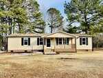 A beige, single-story house with black shutters, a small covered porch, and a brick foundation sits on a large, grassy lot with tall pine trees in the background under a clear sky.