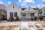 A white wooden house with dark shutters, a covered front porch, and steps leading to the entrance. The yard is partially covered with snow, and some bushes are near the porch. The sky is blue with scattered clouds.