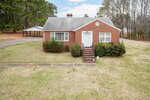 A single-story red brick house with a white door, front steps, and bushes lining the front. The house sits on a grassy lawn with a driveway leading to a detached double carport. Trees surround the property.
