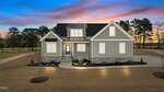 A modern gray house with white trim and stone accents is pictured at dusk, with lights on inside. The home has a covered porch, landscaped front yard, and is surrounded by trees under a colorful sunset sky.