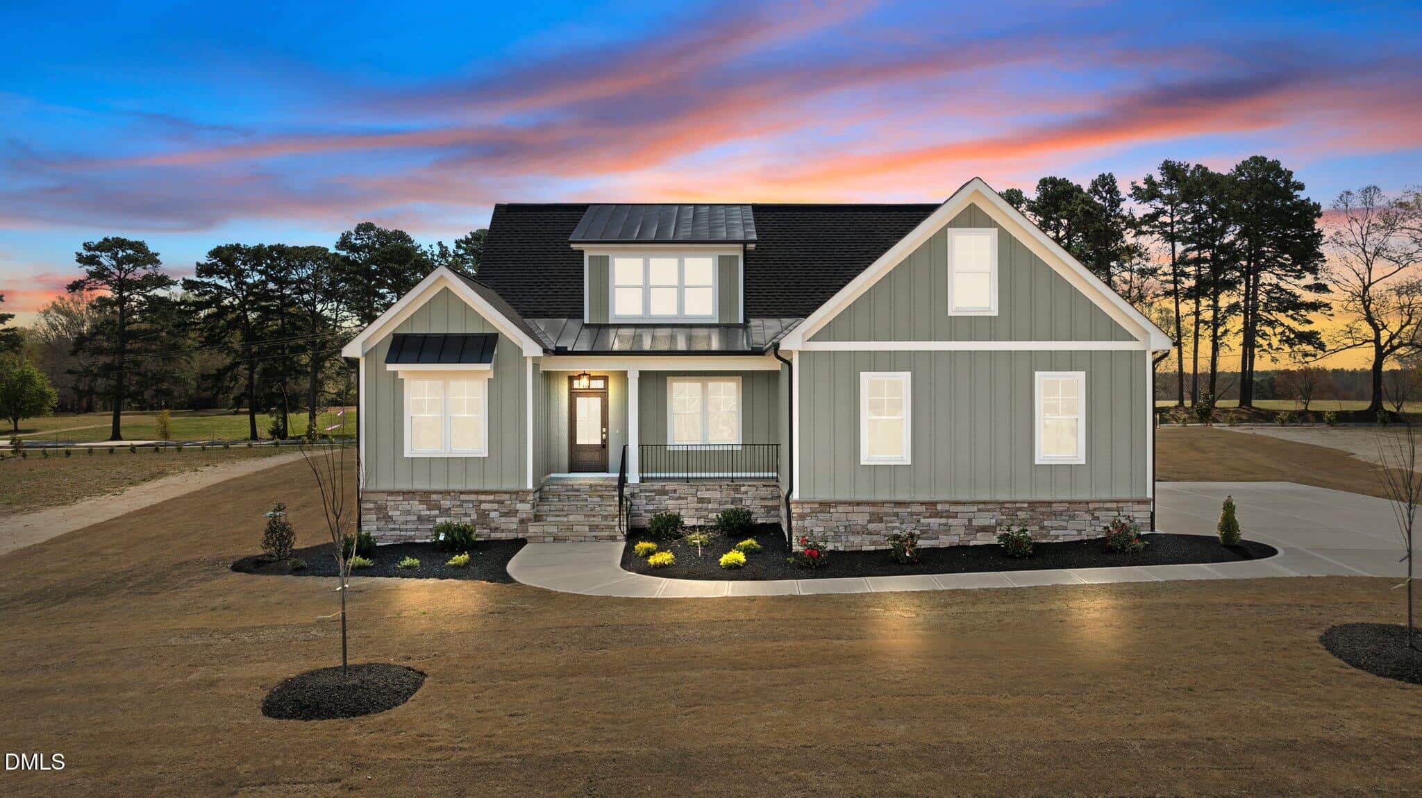 A modern gray house with white trim and stone accents is pictured at dusk, with lights on inside. The home has a covered porch, landscaped front yard, and is surrounded by trees under a colorful sunset sky.