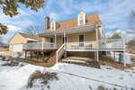 A two-story house with a large front porch, white railings, tan siding, and a brown roof. Snow covers the ground, and a detached garage is visible to the left. Leafless trees and a blue sky are in the background.