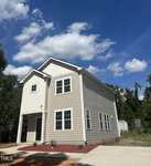 A modern two-story beige house with white trim and multiple windows sits under a partly cloudy blue sky. The front yard features red mulch landscaping and a concrete driveway, surrounded by trees and greenery.