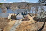 Aerial view of a two-story house with a white exterior and attached garage, situated by a lake with several docks. The yard is surrounded by bare trees and neighboring houses, and a winding driveway leads to the home.