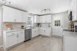 Bright, modern kitchen with white cabinets, stainless steel appliances, light wood flooring, tile backsplash, and a window and door letting in natural light. Minimalistic decor on countertops.