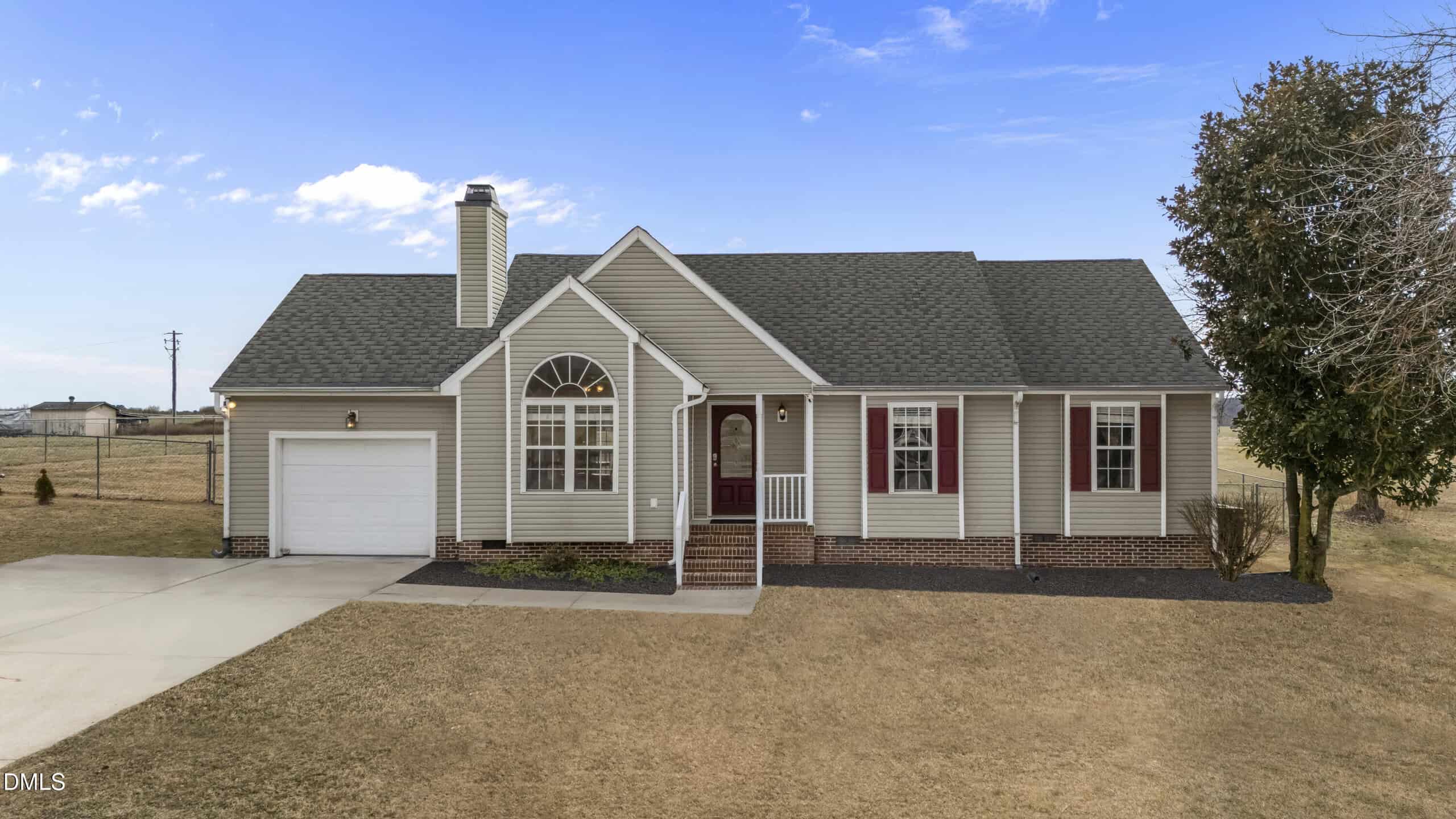 Single-story house with gray siding, brick accents, a front porch, large arched window, and attached single-car garage, set on a lawn with a tree and a clear blue sky in the background.