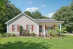 Single-story beige house with red shutters, a covered front porch, and white columns. The yard is neatly landscaped with green grass, shrubs, banana plants, and trees in the background under a blue sky with scattered clouds.