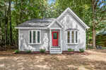 A small, gray house with white trim and a bright red front door, featuring two bay windows, sits in a wooded area with trees and mulch surrounding the front yard.
