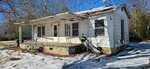 A small, weathered white house with a rusted metal porch roof, metal railings, and a concrete front porch sits on a snowy yard under clear blue skies. The house has peeling paint and overgrown vegetation nearby.