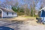 A vacant lot with sparse grass sits between two blue houses on a sunny day. Leafless trees and vines cover the area behind the lot, and two black trash bins are on the right near the curb.