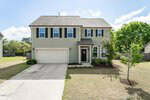 Two-story beige house with black shutters, a double garage, and a covered front porch. The front yard has a small tree, trimmed bushes, and a concrete driveway on a sunny day.