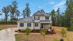 A two-story beige house with a covered front porch, grey roof, and white trim sits on a spacious lot with a curved driveway, young trees, and surrounding tall pine trees under a blue sky.