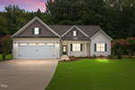 Single-story suburban house with gray siding, a double garage, front porch, and well-maintained lawn, photographed at dusk with exterior lights on and trees in the background.