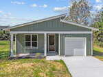 Single-story house with sage green siding, white trim, large front window, and a single-car garage. Concrete driveway and small front yard with new grass and landscaping. Blue sky with scattered clouds in the background.