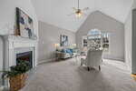 A bright living room with vaulted ceiling, gray carpet, a white fireplace, two armchairs, a blue sofa, abstract art, large windows, plants, and neutral walls. Sunlight streams in through the arched window.