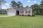 Single-story house with beige siding, white trim, and maroon shutters. Two-car garage, front porch, and flowerbeds with red flowers. Surrounded by green lawn and tall pine trees under a partly cloudy sky.