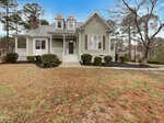 A light green two-story house with white trim, a covered front porch, dormer windows, and a sloped yard with shrubs and pine straw, surrounded by trees under a blue sky.