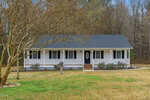 A single-story white house with black shutters and a covered front porch sits on a grassy yard with a large tree in front and a wooded area in the background.