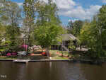 A lakeside house surrounded by tall green trees and colorful flowering bushes, with a small dock extending into the calm water under a partly cloudy sky.