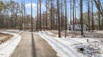 A paved driveway curves through a snowy, wooded area with tall pine trees. In the background, there are RVs and a two-story house surrounded by trees. Sunlight casts long shadows across the snow.