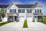 A row of modern townhomes with stone and siding exteriors, blue and light gray doors, white garage doors, small green lawns, and a young tree, all under a bright blue sky.