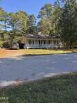 A small, single-story yellow house with a front porch and green shutters sits among tall trees. A gray car is parked in the driveway, and the house faces a quiet, paved road under a clear blue sky.